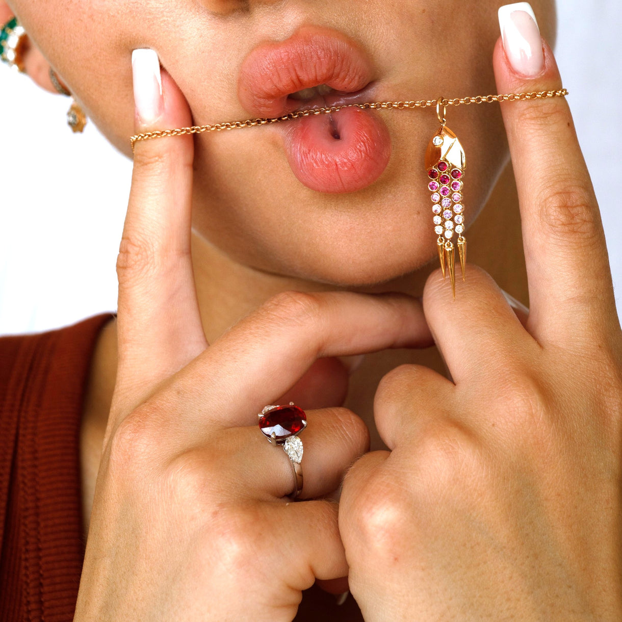 A close-up lifestyle shot of a model wearing a gold chain necklace with a dangling gold pendant decorated with pink and white stones, held across her lips with her hands.