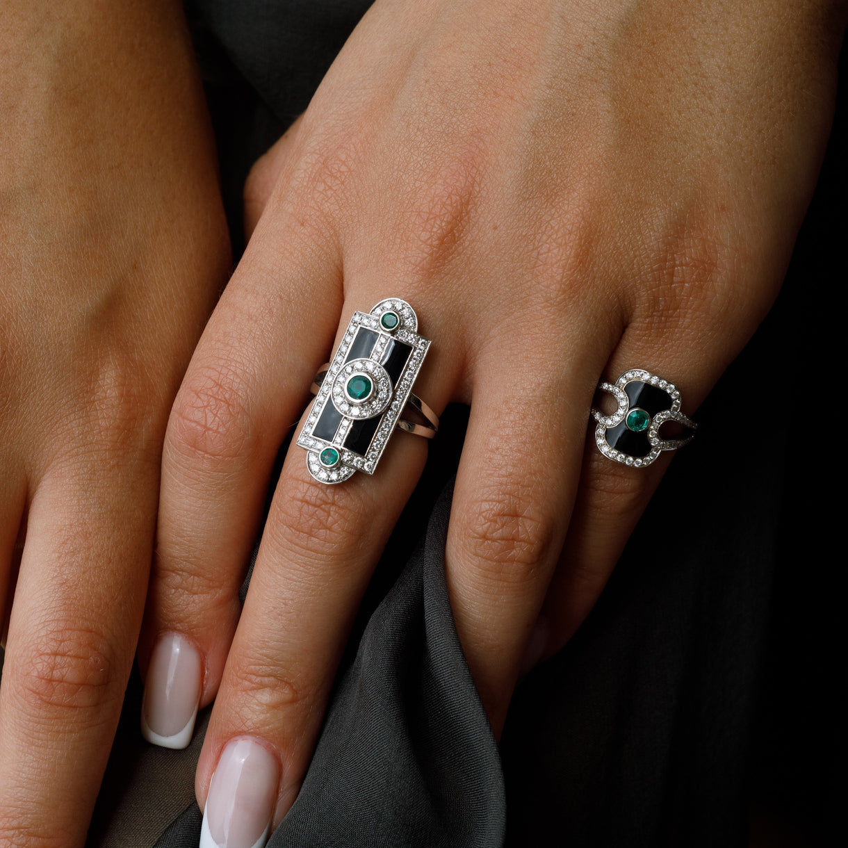 Two Art Deco style rings on a hand. The larger ring on the middle finger features a silver-toned geometric frame with black enamel, a central green stone, and small clear stones.